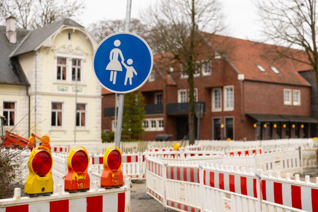 Absperrgitter mit roten Warnleuchten sichern einen Gehweg. Im Vordergrund steht ein blaues Verkehrsschild für einen Fußgängerweg, im Hintergrund sind historische Gebäude zu sehen.